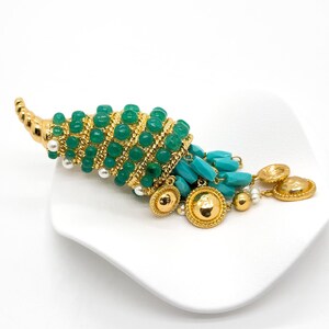 May include: A gold tone brooch with a green and turquoise stone design. The brooch is shaped like a seashell with a long, curled tail. The seashell is covered in green stones and the tail is adorned with turquoise stones and gold beads.