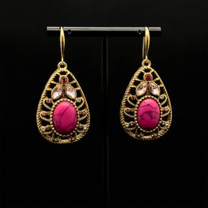 May include: A pair of gold tone dangle earrings with pink and clear stones. The earrings feature a teardrop shape with a pink stone in the center and a filigree design.