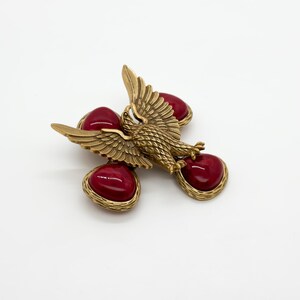 May include: A vintage gold-tone brooch featuring an eagle with outstretched wings, accented by four heart-shaped, deep red cabochons. The brooch has a textured finish and a classic, elegant design, suitable for adding a touch of vintage flair.