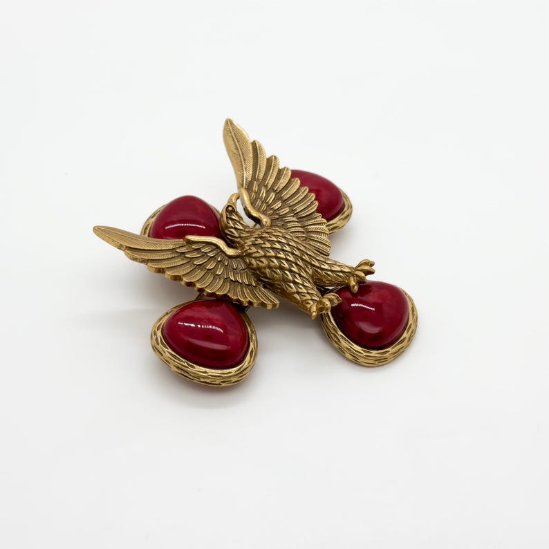 May include: A vintage gold-tone brooch featuring an eagle with outstretched wings, accented by four heart-shaped, deep red cabochons. The brooch has a textured finish and a classic, elegant design, suitable for adding a touch of vintage flair.