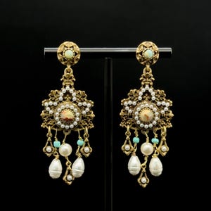 May include: A pair of antique-style gold-coloured earrings. Each earring has a central stone, faux pearls, and turquoise beads. Teardrop-shaped faux pearls dangle from the bottom. The earrings are displayed against a black background.