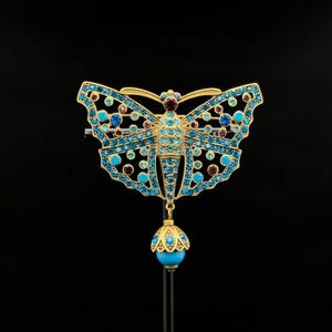May include: A gold-coloured butterfly brooch adorned with turquoise, blue, and burgundy gemstones. The butterfly features openwork wings and a dangling turquoise bead. The brooch is set against a black backdrop.