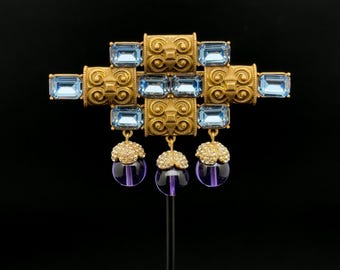 May include: A gold-toned brooch with a geometric design. It features rectangular light blue gemstones and dangling purple and clear orb-shaped pendants. The brooch is displayed against a black background.