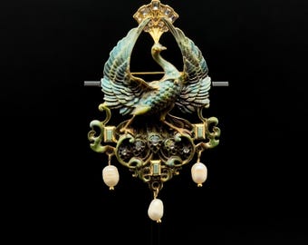 May include: A gold-tone brooch featuring a central bird with outstretched wings, accented with blue and green enamel, clear crystals, and three dangling white pearls. The design includes intricate scrollwork and a fan-shaped top element.