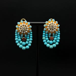 Op de afbeelding: Een paar sierlijke oorbellen met een goudkleurige zetting. De oorbellen hebben een bloemmotief met oranje en witte accenten, omringd door turquoise kralen. Een kleine rode bloem bevindt zich aan de onderkant van de turquoise kralencluster.