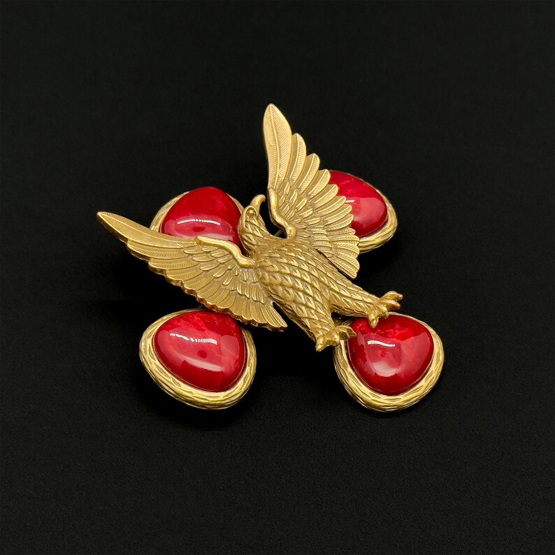 May include: A gold-toned brooch featuring an eagle with outstretched wings, positioned above four heart-shaped red stones. The brooch has a detailed, textured design and is set against a black background.