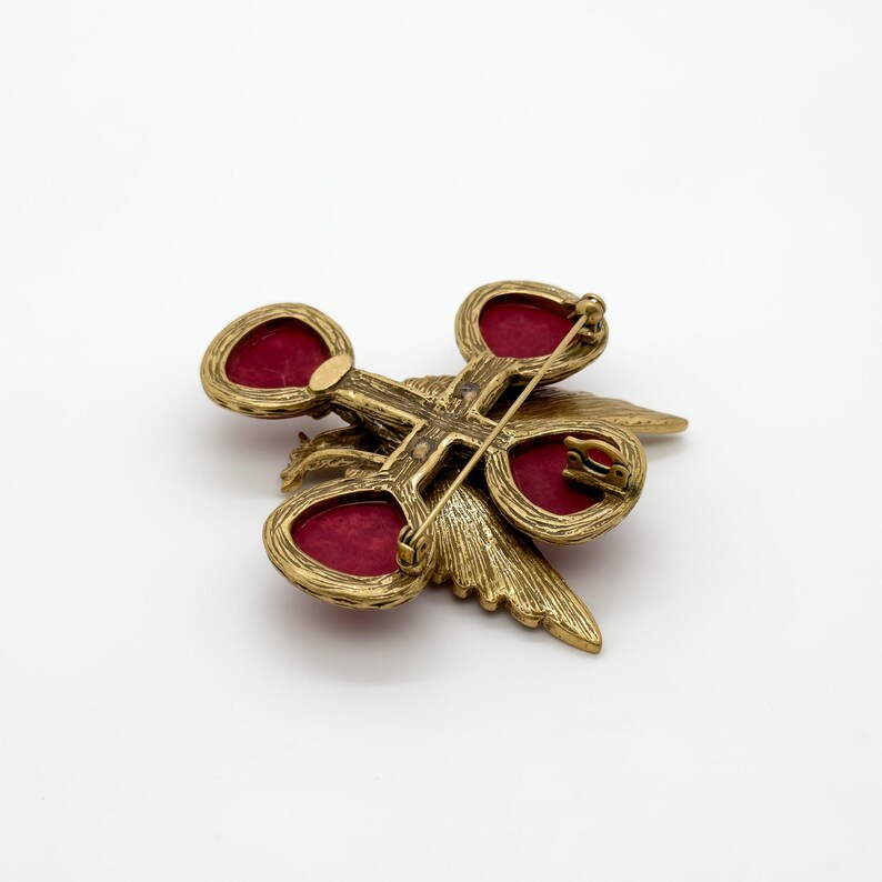 May include: A vintage gold-tone brooch with a cross-shaped design, featuring four oval red cabochons. The brooch has a detailed, textured finish and a pin closure on the back. The brooch is set against a white background.
