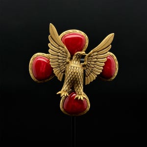 May include: A gold-toned eagle brooch with outstretched wings, perched atop a red, oval-shaped stone. The brooch features four additional red stones, set in gold frames, arranged in a cross shape. The background is black.