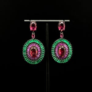 May include: A pair of drop earrings with a striking design. Each earring has a large, oval, ruby-red central stone, encircled by concentric rings of green and pink stones. The earrings are suspended from a smaller ruby-red stone.