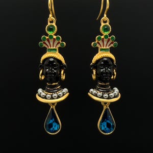 以下が含まれることがあります： 黒のエナメルで顔と緑と白の冠が施された金のイヤリング。イヤリングには、下から青い涙型の宝石がぶら下がっています。