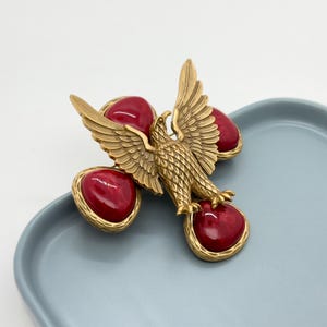 May include: A gold-toned eagle brooch with outstretched wings and a textured body. The eagle is surrounded by four heart-shaped, deep red cabochons set in gold-toned frames. The brooch is displayed on a light blue surface.