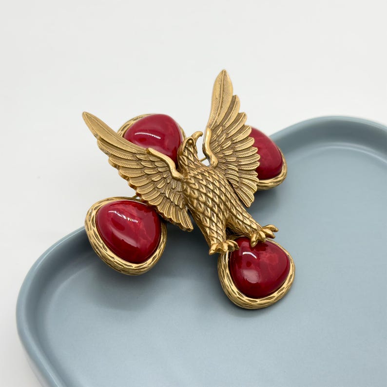 May include: A gold-toned eagle brooch with outstretched wings and a textured body. The eagle is surrounded by four heart-shaped, deep red cabochons set in gold-toned frames. The brooch is displayed on a light blue surface.