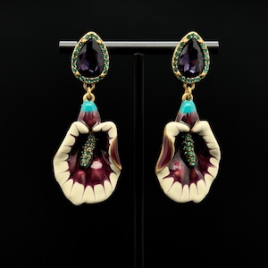 May include: A pair of gold-toned earrings featuring a calla lily design. Each earring has a purple teardrop-shaped gem surrounded by green stones, a turquoise accent, and a cream, burgundy, and green calla lily pendant.