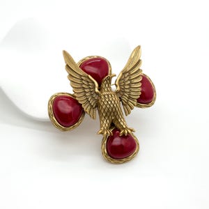 May include: A vintage gold-tone metal eagle brooch with outstretched wings, perched above four large, oval, deep red cabochon stones. The eagle has detailed feathering. The brooch is set against a white background.