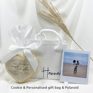 May include: A heart-shaped biscuit, wrapped in cellophane and tied with a white ribbon, has the text "Will You Be My Godmother". A white gift bag with "Hannah" and a Polaroid photo are also in the image.