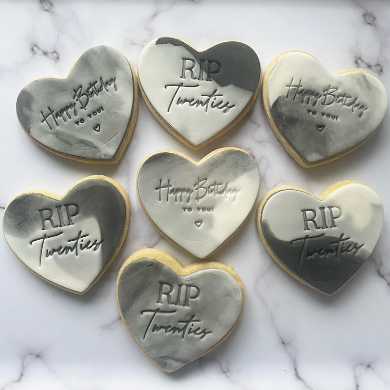May include: Seven heart-shaped cookies decorated with gray and white marbling. The cookies have different messages, including "Happy Birthday to You!" and "RIP Twenties".
