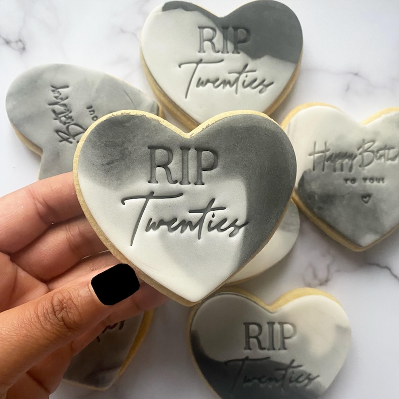 May include: Four heart-shaped cookies decorated with gray and white marbling. The cookies have the text "RIP Twenties" in gray icing.