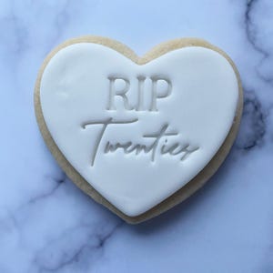 May include: A white heart-shaped cookie with the text "RIP Twenties" in black lettering.