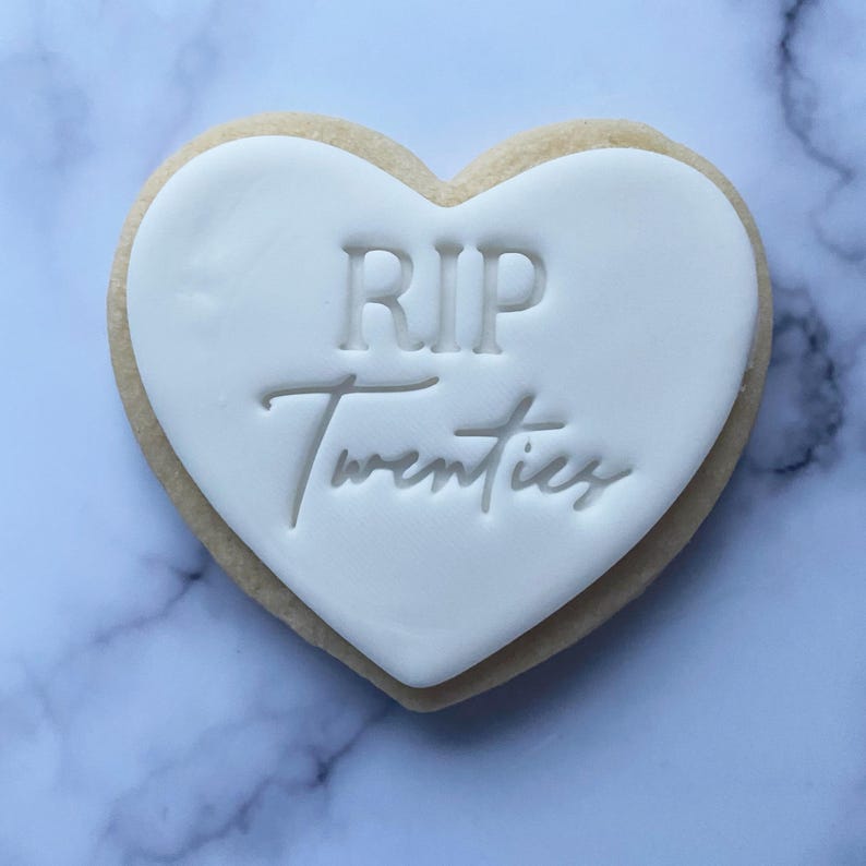 May include: A white heart-shaped cookie with the text "RIP Twenties" in black lettering.