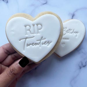 May include: Two heart-shaped sugar cookies decorated with white icing. One cookie says "RIP Twenties" and the other says "Happy Birthday to you".