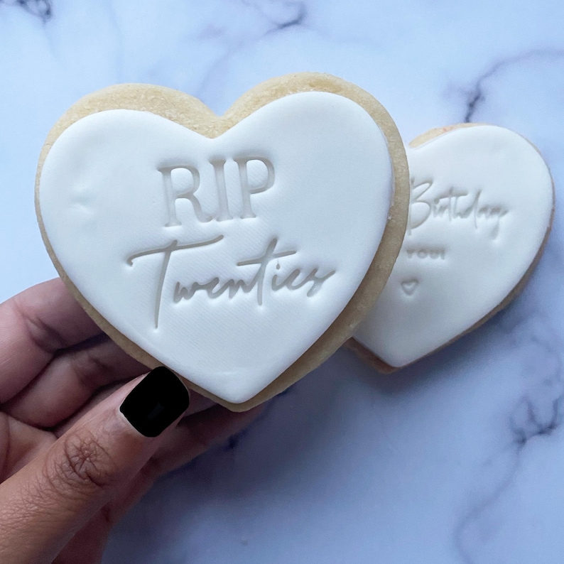 May include: Two heart-shaped sugar cookies decorated with white icing. One cookie says "RIP Twenties" and the other says "Happy Birthday to you".