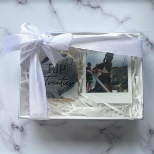 May include: A white gift box with a white ribbon and bow. The box contains a cookie decorated with a tombstone and the text "RIP Twenties" and a photo of a person on a boat.