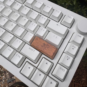 May include: A white mechanical computer keyboard featuring a rectangular wooden keycap with a visible grain pattern replacing the standard right shift key. The keyboard includes standard alphanumeric keys, a rotary knob, and function keys.