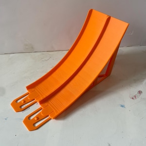 May include: Bright orange toy car track ramp. The ramp has two parallel lanes and a support structure. The track is made of plastic and is designed for toy cars. The ramp is angled upwards.