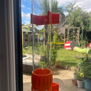 May include: A clear, cylindrical tube mounted vertically against a glass surface using two orange plastic brackets. Each bracket is secured to the glass with a transparent suction cup, holding the tube firmly in place for display or utility.