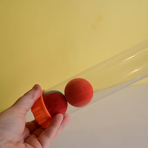 Op de afbeelding: Een hand houdt een oranje plastic beker met twee rode ballen vast. Een doorzichtige plastic buis is naar rechts gekanteld, met een rode bal erin. De achtergrond is een effen gele muur.