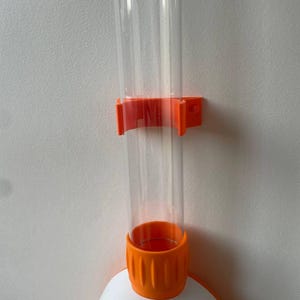 Könnte beinhalten: Ein klares Kunststoffrohr wird von einer orangefarbenen Halterung an einer weißen Wand gehalten. Das Rohr ist mit einem weiß-orangenen Sockel verbunden. Der Sockel hat eine orangefarbene, geriffelte, zylindrische Komponente.
