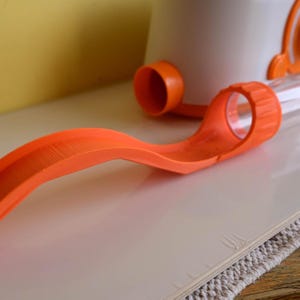 Op de afbeelding: Een oranje plastic speelgoedbaan met een doorzichtige buis en een wit-oranje container. De baan is gebogen en heeft een gestructureerd oppervlak. Het speelgoed ligt op een wit oppervlak.