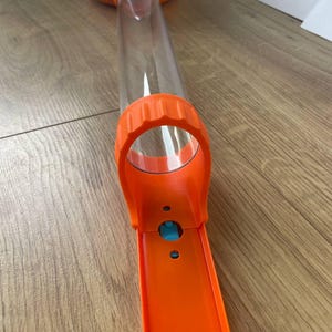 Könnte beinhalten: Ein orange-weißes Spielzeug mit einem durchsichtigen Kunststoffrohr und einem blauen Knopf. Das Spielzeug hat eine lange, orangefarbene Bahn und einen weißen Sockel mit orangefarbenen Akzenten. Das transparente Rohr ist mit der orangefarbenen Bahn verbunden.