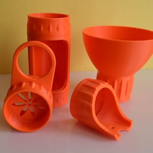 May include: Orange 3D-printed objects, including a cylindrical container with a handle, a funnel-shaped piece, and two other components. The objects are arranged on a white surface against a yellow background. The components appear to be parts of a larger device.