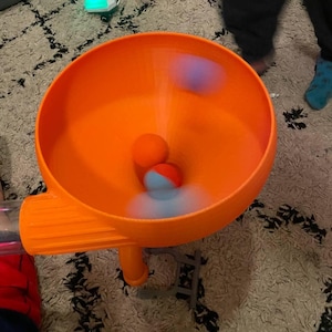 May include: An orange plastic toy with a funnel shape and a tube. Several colourful balls are inside the funnel, appearing to be in motion. The toy is on a patterned rug.