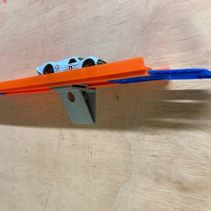 May include: A blue toy car is driving on an orange and blue track. The track is supported by two grey brackets. The track is mounted on a wooden surface.