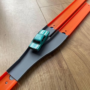 May include: A green toy car on a gray and orange plastic track set. The track is designed to create a split path for the car to travel.