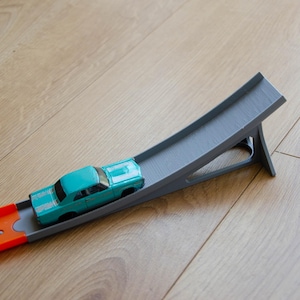 May include: A blue toy car drives up a grey plastic ramp with an orange track at the bottom.