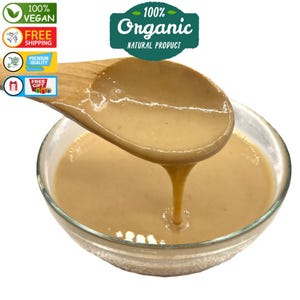 May include: A wooden spoon scoops a light brown, creamy substance from a glass bowl. The bowl is filled with the same creamy substance. The text "100% Organic Natural Product" is visible in the image.