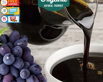 May include: A close-up of a dark, thick liquid being poured from a glass jug into a white bowl. The liquid is grape molasses, a natural product made from grapes. A bunch of dark purple grapes is in the foreground.