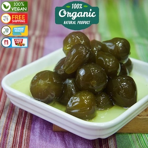 Può includere: Un piatto quadrato bianco pieno di confettura di fichi verdi. Il piatto è su una superficie a strisce colorate. Il testo "100% Organic Natural Product" è visibile nell'angolo in alto a destra dell'immagine.