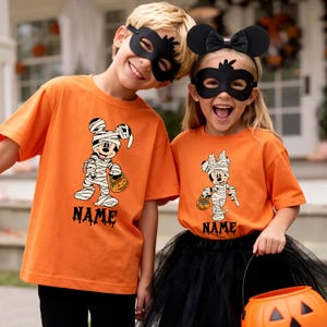 May include: Two orange short-sleeve t-shirts featuring a graphic of a mummy-wrapped cartoon mouse holding a pumpkin bucket. Below the graphic, the word NAME is printed in a black, dripping font. The shirts are worn with black masks.
