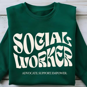May include: A dark green sweatshirt with the words "SOCIAL WORKER" in a white retro font. The text "ADVOCATE. SUPPORT. EMPOWER." is printed below the main text.