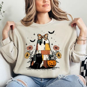 May include: Beige sweatshirt featuring a Halloween-themed graphic. The design includes a patchwork ghost with a bow, a black cat, a jack-o'-lantern, flowers, and bats. The sweatshirt is a comfortable, casual garment for the fall season.