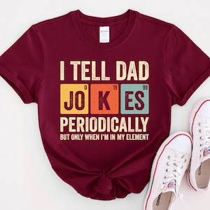 May include: A maroon t-shirt with the text "I TELL DAD JOKES PERIODICALLY BUT ONLY WHEN I'M IN MY ELEMENT" in a colorful, periodic table-inspired design. The shirt is paired with white sneakers.