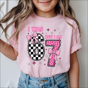 May include: Pink t-shirt with the text "I WAS NOW I AM 67". The number 6 is black and white checkered, and the number 7 is pink leopard print. Both numbers have pink bows and star accents.