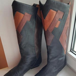 May include: A pair of knee-high boots featuring a black base with geometric panels in shades of tan and dark brown. The boots have a pointed toe, a flat sole, and a V-shaped cutout at the top of the shaft, showcasing a multi-toned leather construction.