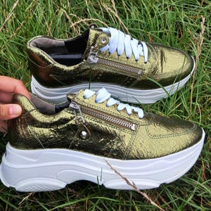 May include: A pair of metallic olive green sneakers with a textured, cracked leather-like finish. These shoes feature white laces, a prominent side zipper detail, and a thick, chunky white rubber platform sole with a textured tread pattern.