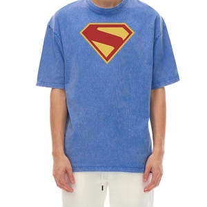 Peut inclure: Un t-shirt bleu à manches courtes avec une texture délavée, arborant un emblème en forme de losange jaune et rouge sur la poitrine. Le motif se compose d'une lettre S stylisée dans un champ jaune avec une bordure rouge, centré sur le devant.