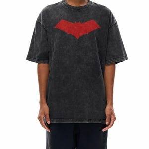 Puede incluir: Camiseta negra con un aspecto desgastado, con un gran símbolo de murciélago rojo en la parte delantera. La camiseta tiene mangas cortas y cuello redondo. El símbolo del murciélago es un gráfico llamativo.